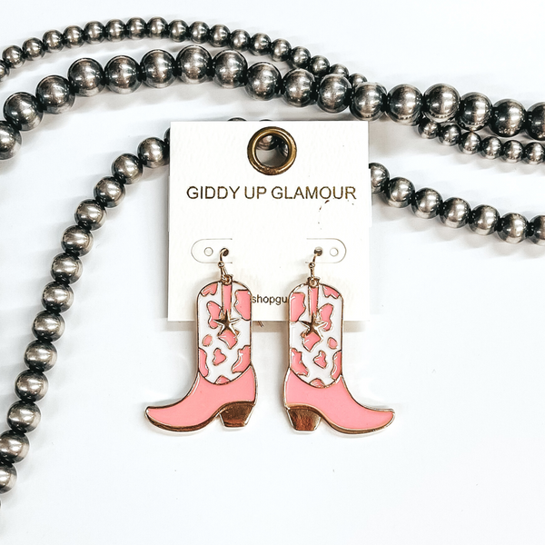 Gold fish hook earrings with a hanging boot pendant. This boot has a pink and white cow print with a small gold star. These earrings are pictured on a white background with silver beads. 