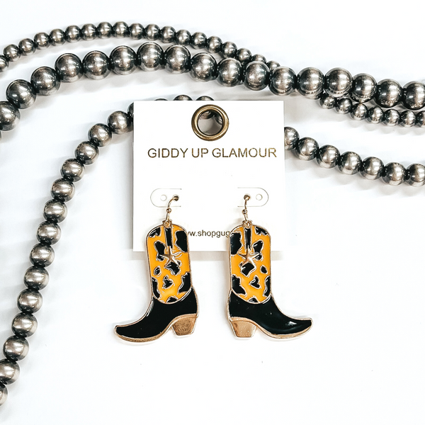 Gold fish hook earrings with a hanging boot pendant. This boot has a brown and black cow print with a small gold star. These earrings are pictured on a white background with silver beads. 