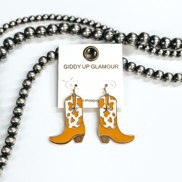 Gold fish hook earrings with a hanging boot pendant. This boot has a tan and ivory cow print with a small gold star. These earrings are pictured on a white background with silver beads. 