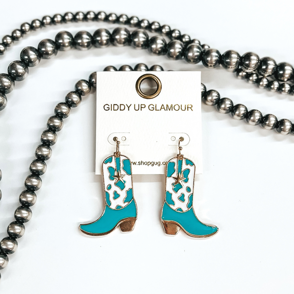 Gold fish hook earrings with a hanging boot pendant. This boot has a turquoise and white cow print with a small gold star. These earrings are pictured on a white background with silver beads. 
