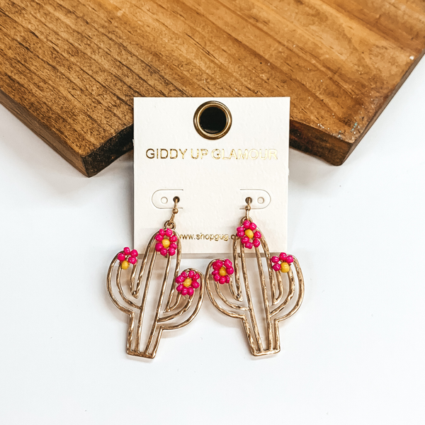 Gold fish hook earrings with a hanging gold cactus outline pendant. the cactus pendant also has pink beaded flowers at the top. These earrings are pictured in front of a wood block on a white background. 