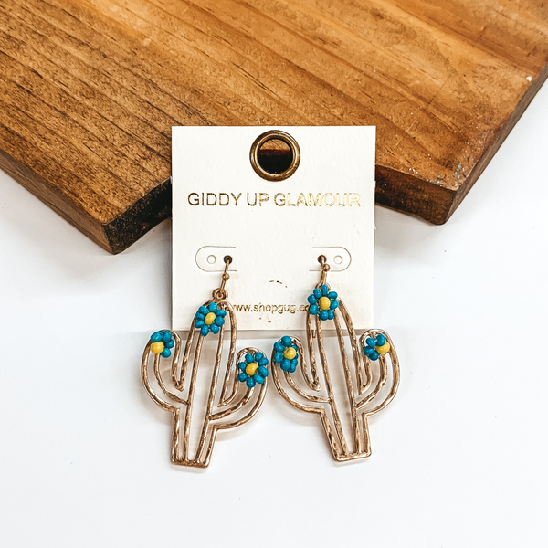 Gold fish hook earrings with a hanging gold cactus outline pendant. The cactus pendant also has blue beaded flowers at the top. These earrings are pictured in front of a wood block on a white background.