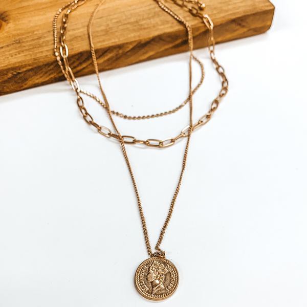 Three layered gold chain necklace with a double sided coin. Each strand is a different length with a different style chain. The longest stran dhas the coin. This necklace is pictured on a white background while partially laying on a brown block. 