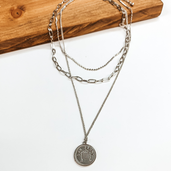 Three layered silver chain necklace with a double sided coin. Each strand is a different length with a different style chain. The longest stran dhas the coin. This necklace is pictured on a white background while partially laying on a brown block. 