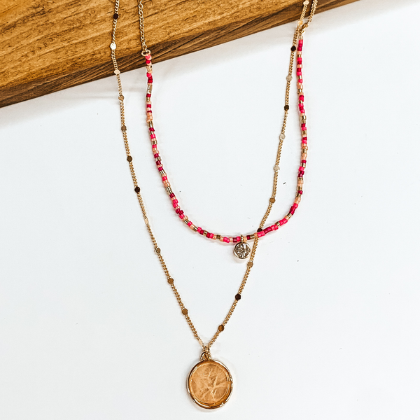 Two stranded necklace. The shortest strand has pink colored beads with a clear crystal charm. The longest strand is a gold chain with a taupe colored charm. This necklace is pictured on a white background while partially laying on a brown block. 