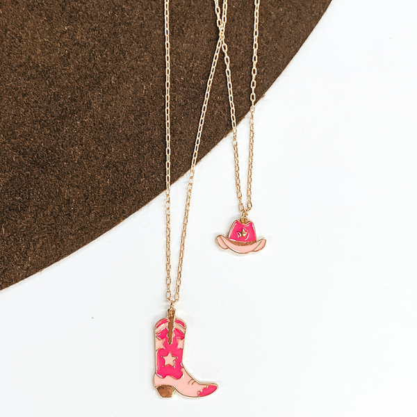Two stranded necklace. The shortest strand is a gold chain with a light pink and dark pink hat charm. The longest strand is a gold chain with a light pink and dark pink star print boot charm. This necklace is pictured on a white and brown background.  