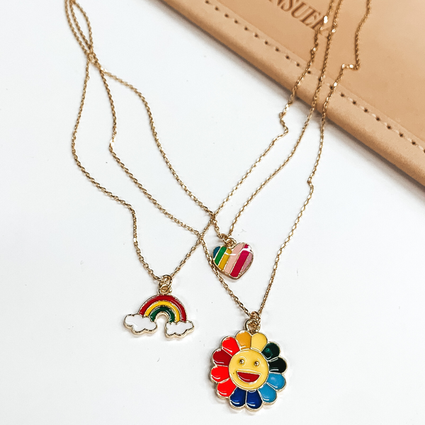 Three stranded gold chain necklace. Each strand has an individual charm multicolored. These include, a heart charm, a rainbow charm, and a happy face flower charm. This necklace is pictured partially on a tan bag on a white background. 