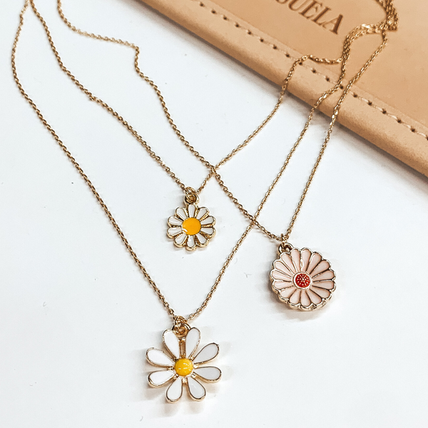 Three stranded gold chain necklace. Each strand has an individual charm in colors white, yellow, and pale pink. These include, two white flower charms in different sizes and a pale pink flower charm. This necklace is pictured partially on a tan bag on a white background. 