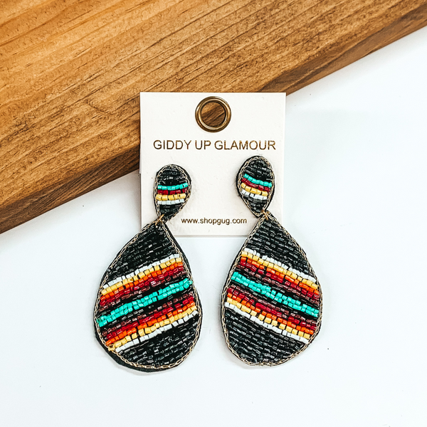 Beaded upside down teardrop stud earrings with a hanging teardrop. The main color of the beads is black with multicolored stripes. These earrings are pictured on a white and brown wood background.
