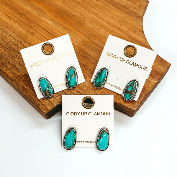 Oval turquoise stone earrings with a silver outline. There are three pairs of studs all on white earring holders. Two pairs are pictured on a wood block laying on a white background and the third is pictured on a plain white background.