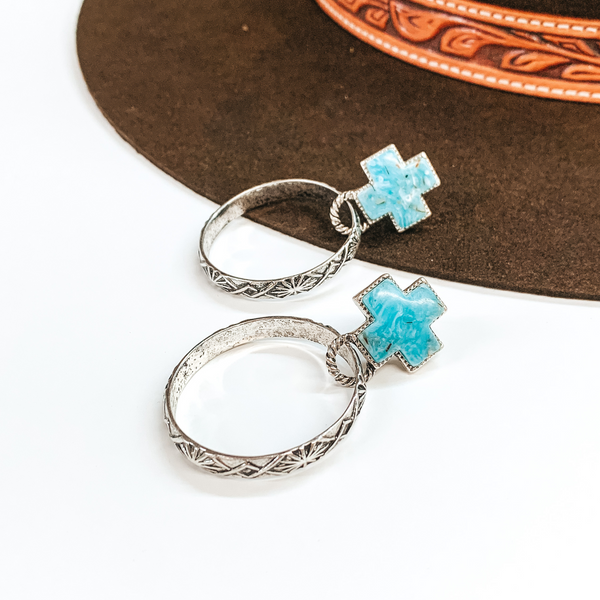 Turquoise cross studs with a silver outlining. These earrigs also have silver, open drop circles with an engraved design. One earrings is partially laying on a brown hat on a white background while the other earrings is fully on the white background. 