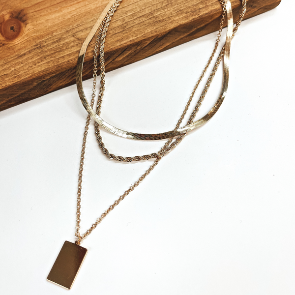 Gold chain necklaces pictured partially laying on a wood block with the rest of the necklaces on a white background. This pictures includes a snake chain, a longer rope chain, and an even long regular chain with a gold rectangle pendant.