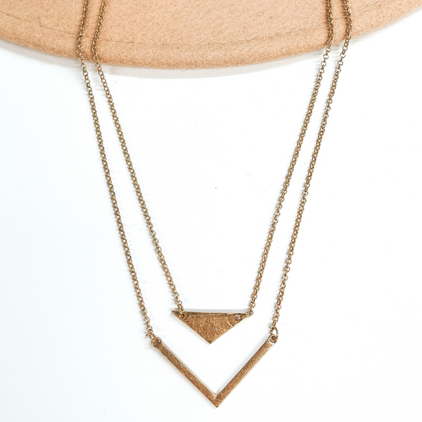 Gold, two strand necklace pictured on a white background. The shorter strand has a wide triangle pendant, while the longer strand has a thing arrow head. 