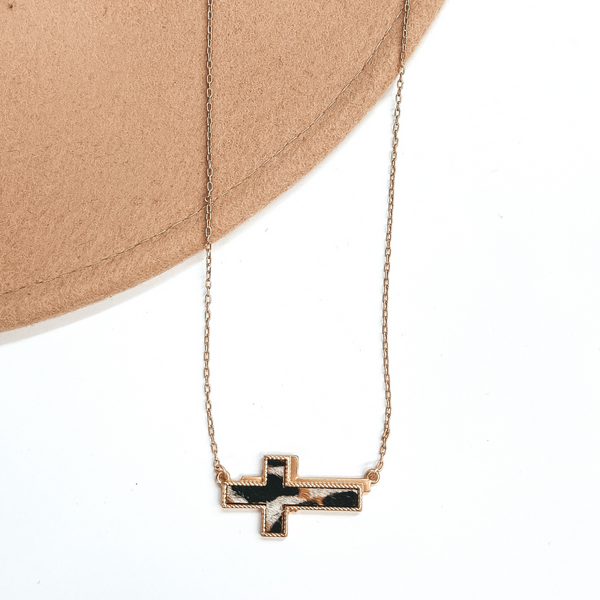Gold chain necklace with a gold cross pendant with a leopard print inlay. This necklace is pictured on a brown and beige hat. 