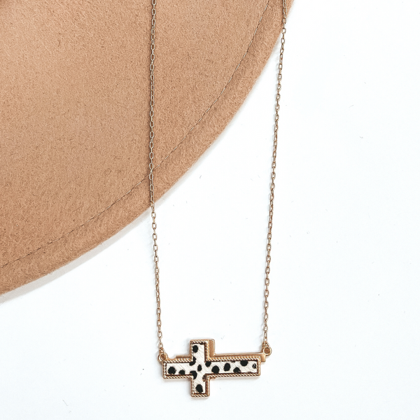 Gold chain necklace with a gold cross pendant with a dotted print inlay. This necklace is pictured on a brown and beige hat. 