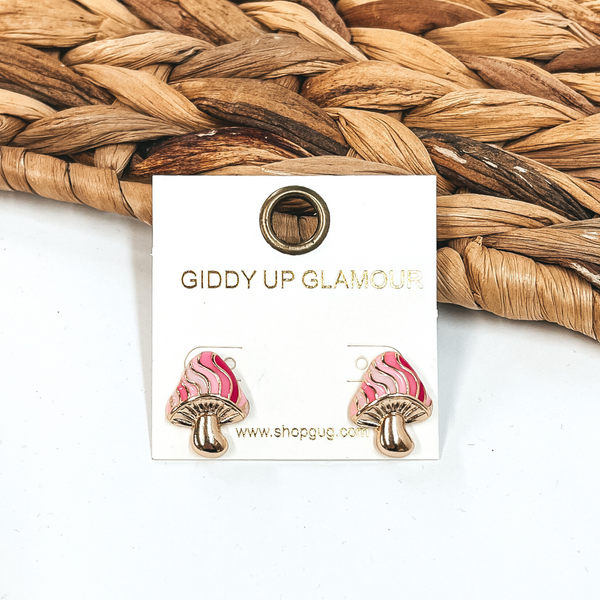 Gold, mushroom shaped earrings. The top part of the mushroom has different shades of pink stripes. These earrings are pictured in front of a basket weave material on a white background. 