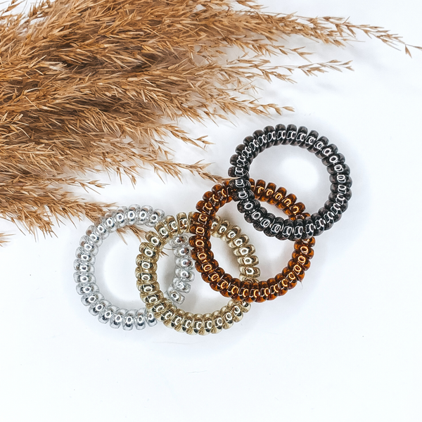 Pictured are four spiral scrunchies laying on top of each other in the colors black, brown, gold, and silver. These scrunchies are pictured on a white background with tan pompus grass in the top left corner.