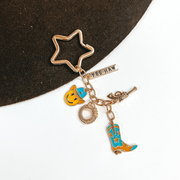 Gold, star shaped key ring with a hanging gold chain with charms on it. There is a gold rectangle charm with the words "YEE HAW," a gold gun charm, a gold rope charm, a yellow smiley face chram with a turquoise cowboy hat, and a turquoise and tan cowboy boot charm. This is pictured on a brown and white background. 