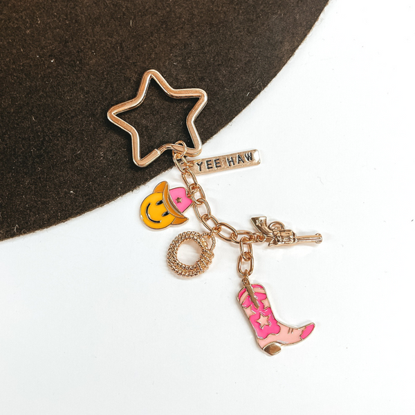 Gold, star shaped key ring with a hanging gold chain with charms on it. There is a gold rectangle charm with the words "YEE HAW," a gold gun charm, a gold rope charm, a yellow smiley face chram with a pink cowboy hat, and an light pink and pink cowboy boot charm. This is pictured on a brown and white background. 
