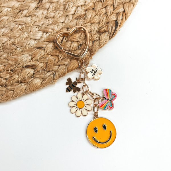 Gold, heart shaped key ring with a hanging gold chain with charms on it. There is a gold flower charm with the words "Be Happy," a gold bumble bee charm, a white flower charm, a rainbow butterfly charm, and a yellow smiley face charm. This is pictured partially on a basket weave material on a white background. 
