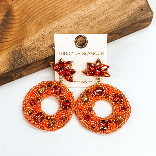 Half flower red crystal stud earrings with an orange beaded wreath hanging from the bottom of the studs. These earrings are pictured in front of a wood block on a white background. 