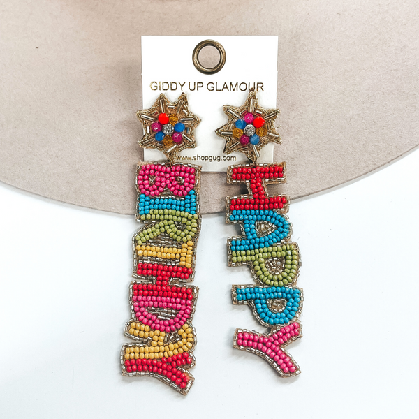 These are multicolor seedbeaded earrings with the saying, 'Happy Birthday'. The post back has a gold ribbon with small crystal beads in the center and a clear crystal. The right earrings says, 'Happy', with seedbeads in  red, blue, green, and pink with a gold outline. The left earring says, 'Birthday', in colors pink, blue, green, yellow, and red. These earrings are taken  laying on a beige hat and a white background.
