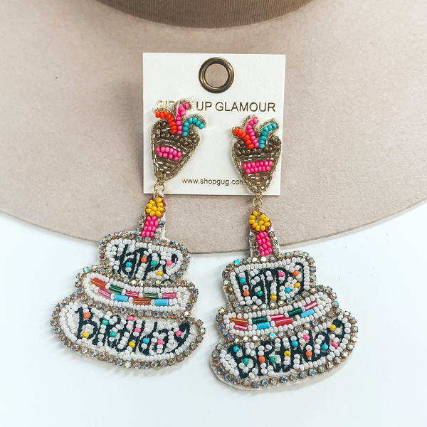 Three tiered birthday cake earrings with seedbeads in white with the  saying, 'Happy Birthday', in black. The middle tier has long crystal  beads in various colors. The cake is surrounded by ab crystals all around  and there is one pink candle on the top with a yellow tip. The postback  part has a gold beaded confetti holder with confetti in colors turquoise,  orange, and pink. These earrings are taken on a beige hat and on a white  background.