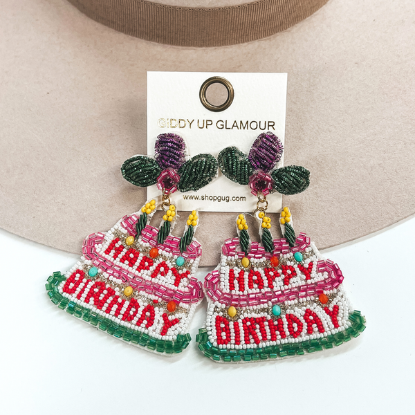 Two tiered birthday cake earrings with the saying, 'Happy Birthday', in  the center. These earrings have three candles on top of the cakes with a  bow postback. The postback part has bows in colors green and purple with  a pink center. The top tier has a pink border and the saying, 'Happy', in red seedbeads  and three candles in green with a yellow tip. The bottom tier has the saying,  'Birthday' with a green border. These earrings are taken on a beige hat  and on a white background.