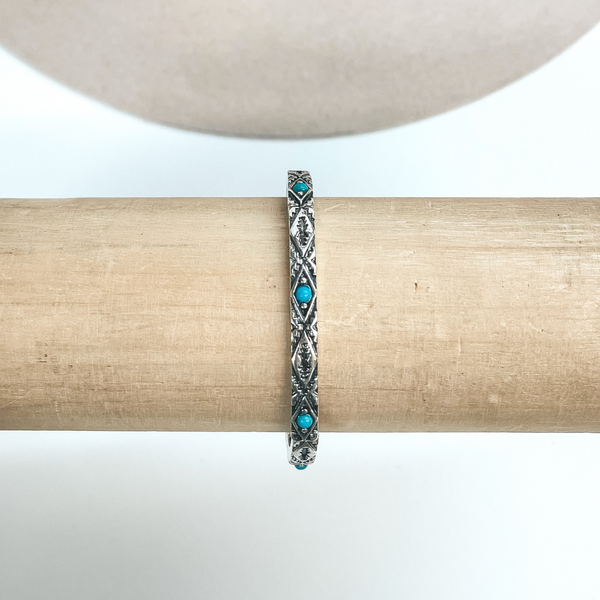 This is a thin silver bracelet with Aztec engraved design and small  turquoise stones all around in the center. These earrings are taken on a  light brown bracelet holder and on  a white background with a beige hat in the back as decor.