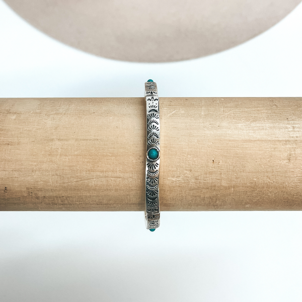 This is a thin silver bracelet with sun engraved designs and small  turquoise stones all around in the center. These earrings are taken on a  light brown bracelet holder and on  a white background with a beige hat in the back as decor.