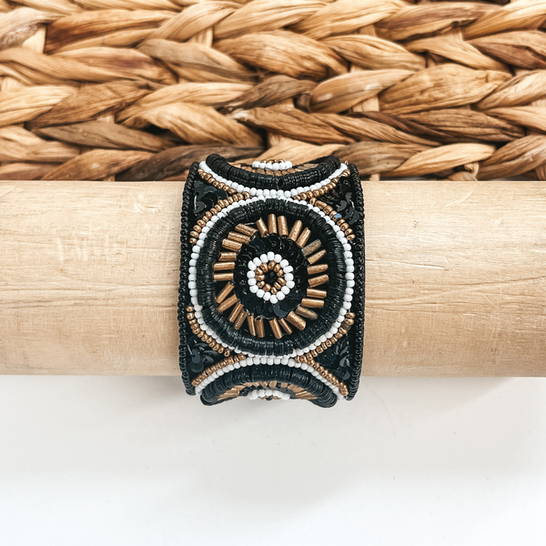This is a beaded bracelet in black, white, and gold with sequins.  There is a variety of different shapes of beads, all in a circle.  This bracelet is taken on a light brown bracelet holder  and on a white background with a brown woven slate in the back as decor.