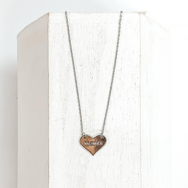 Dainty silver chained necklace with a silver heart pendant. In the center of the pendant has the word "mama" engraved. This necklace is pictured laying on a white block on a white background.