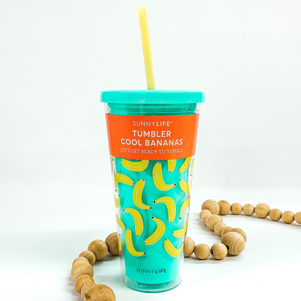 Sky blue double insulated cup. On the outside of the cup there are bright yellow bananas on the cup. This cup includes a yellow straw. This cup is pictured with an orange sticker around the top of the cup with details of the cup. this cup is pictured on a white background with tan beads around it. 