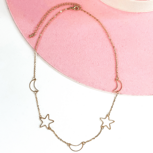 Simple, adjustable, gold chained necklace with three moon charms and two star charms spaced through the bottom half of the necklace. This necklace is pictured on a white and pink background. 