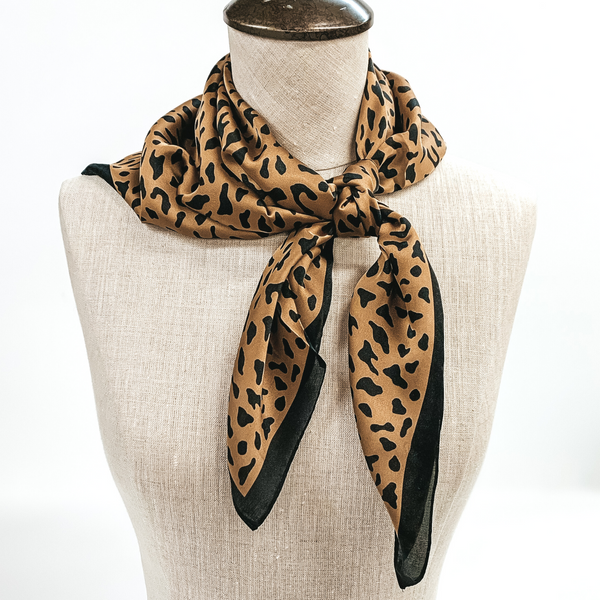Tan scarf with black leopard print and black outline tied around the neck of a mannequin. Pictured on white background.