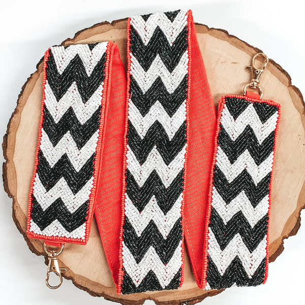 This purse strap has a black and white chevron striped pattern with a coral colored backing. This purse strap has gold clasps. This purse strap is pictured on a piece of wood on a white background. 