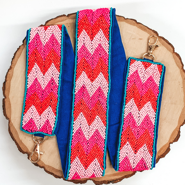 This purse strap has a dark pink, light pink, and orange chevron striped pattern with a navy colored backing. This purse strap has gold clasps. This purse strap is pictured on a piece of wood on a white background.