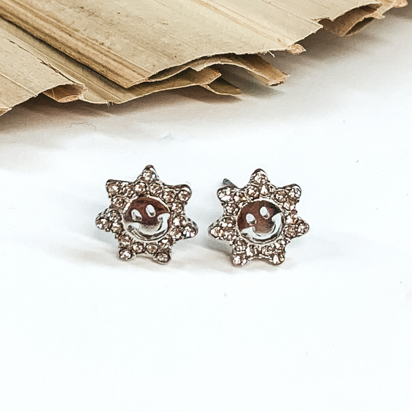 Silver sun shaped stud earrings with a cut out smiley face and clear crystals. These earrings are pictured laying on a green palm leaf on a white background.