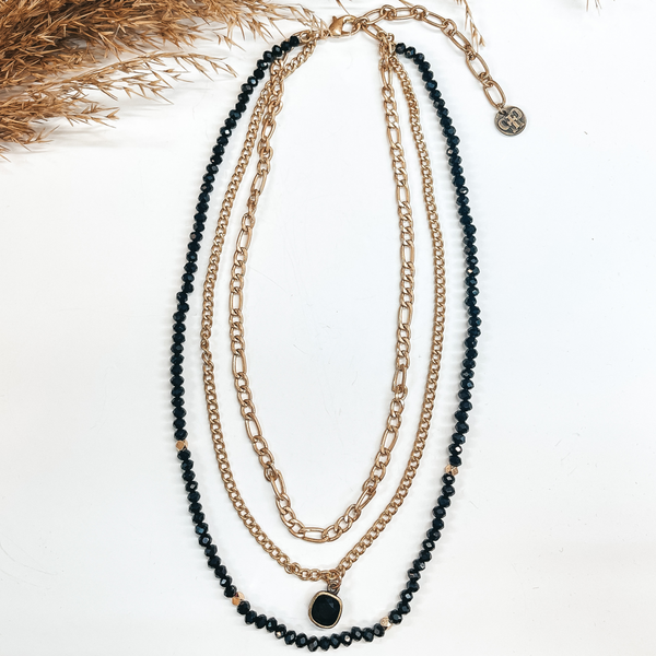 Three strand necklace with a black cushion cut  crystal hanging pendant. The outer strand is all black beads with a few gold spacers. The second  strand is a matte gold chain with the cushion cut crystal. The smaller strand is a matte gold chain. Taken on a white background with a brown plant in the back as decor.