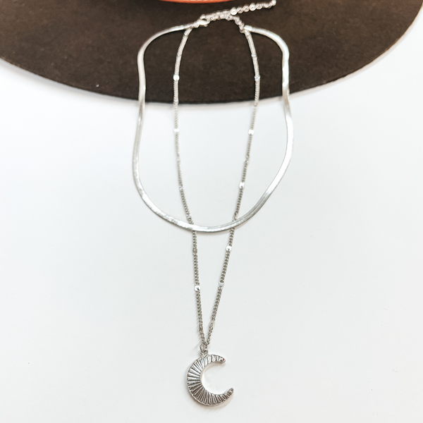 Double layered silver necklace, the smaller necklace  is a snake chain and the bigger one is just a  regular chain. The longer chain has a sunburst  textured moon pendant. Taken on a white background  and laying on a dark brown hat.