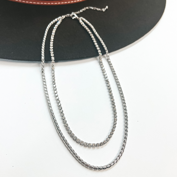 This is double layered silver chain necklace, the longer  chain is a braid textured chain. The smallest chain  has rhinestones all around. This necklace is pictured  laying on a dark brown hat and white background.
