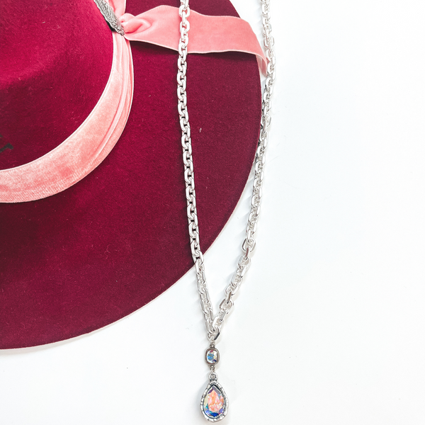 This is a long, thick link chain necklace in silver  with an AB cushion cut crystal connected to an AB  crystal teardrop. This necklace is taken laying on  a burgundy colored Charlie Horse hat and white  background.