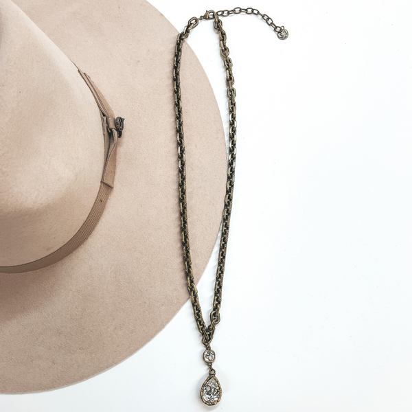 This is a long, thick link chain necklace in  bronze with clear cushion cut crystal connected  to a clear crystal teardrop. This necklace is taken  laying on a light brown hat and white background.
