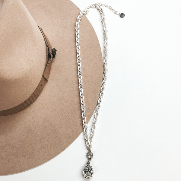 This is a long, thick link chain necklace in  silver with clear cushion cut crystal connected  to a clear crystal teardrop. This necklace is taken  laying on a light brown hat and white background.