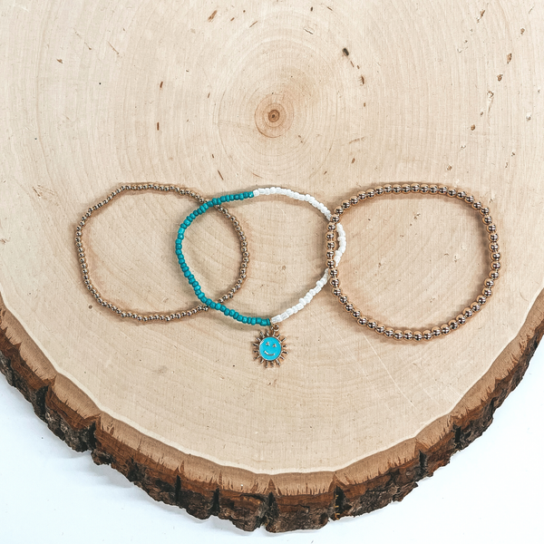 These are three beaded bracelets, the left bracelet   has small gold beads and the right bracelet  has bigger gold beads. The middle bracelet has  turquoise and ivory beads with a turquoise sun  pendant with a happy face. These bracelets are taken  laying on a slab of wood and a white background.