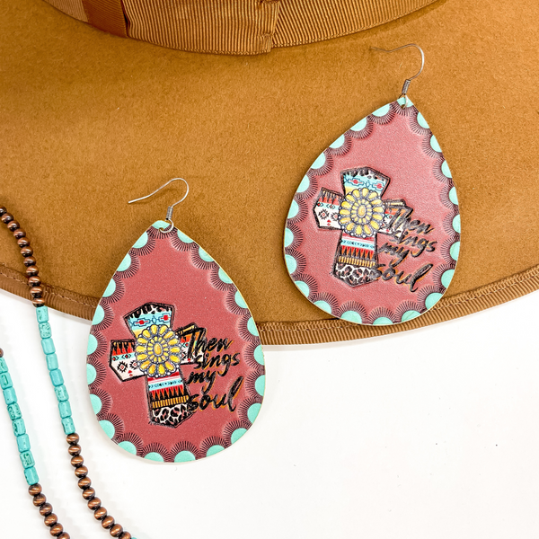 These are brown leather teardrop earrings with the  saying "Then sings my soul" and a aztec prink cross  with a yellow concho in the center. There are teal  green bumps all aorund with black rays. These  earrings are taken on a brown hat brim and a white  background, there are  copper and turquoise beads in the side as a decor.