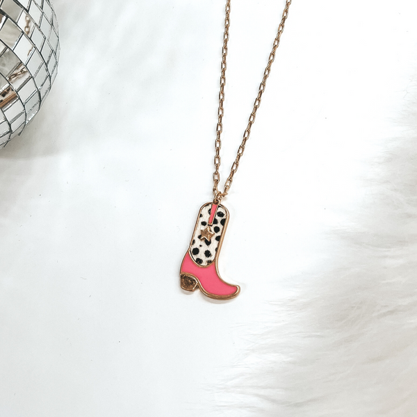 This is a gold chain necklace with a boot pendant in a gold setting. The boot pendant is pink and has a dotted print with a gold star. This necklace is taken on laying on a white background with a white fur carpet in the side and two disco ball as decor.