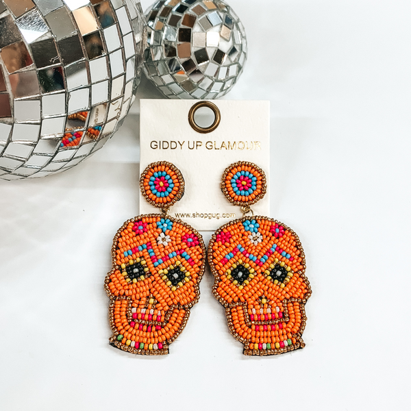 These are seedbeaded sugar skull post earrings in  orange with multicolored beads as well. These earrings  are taken on a white background and two disco balls  in the back as decor.