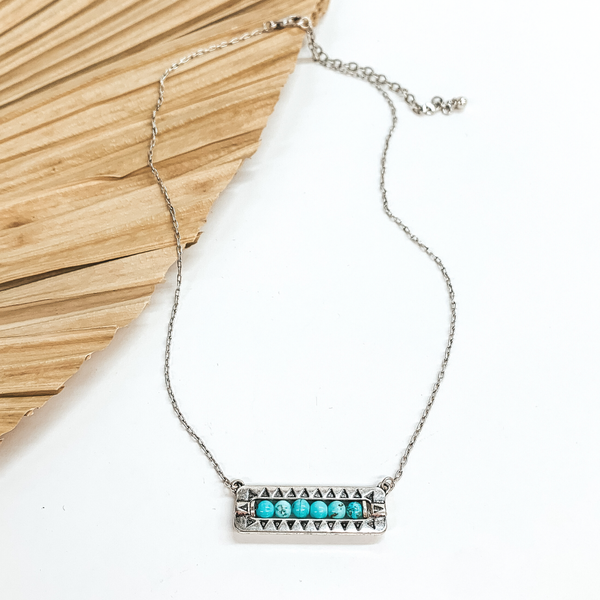 Silver necklace with a horizontal bar pendant with stone beads in  turquoise and black detailing. This necklace is taken laying on a  dried up palm leaf and white background. 