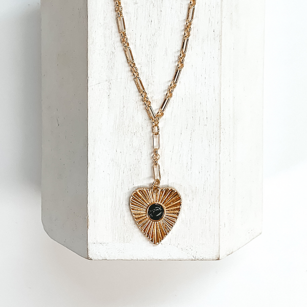 Gold linked chain with a sunburst heart pendant with a small black stone in the center. This necklace is taken on a white block and on a white background.